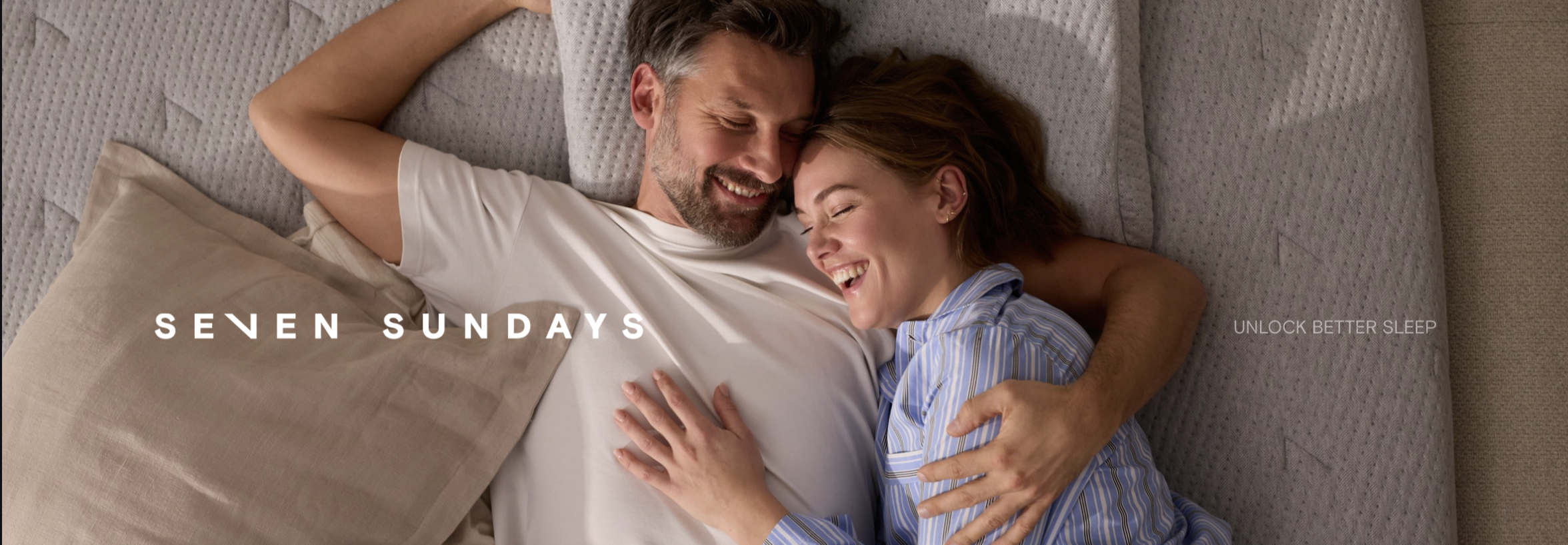 Paar liegt entspannt im Bett und umarmt sich, Kissen mit Seven-Sundays-Logo im Hintergrund